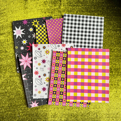 A fan arrangement of eight colorful patterned sticker paper sheets on a textured yellow-green surface. The designs include black and white gingham, pink and yellow gingham, black and yellow check, pink polka dots, white with pink stars and flowers, and a multicolor geometric pattern.