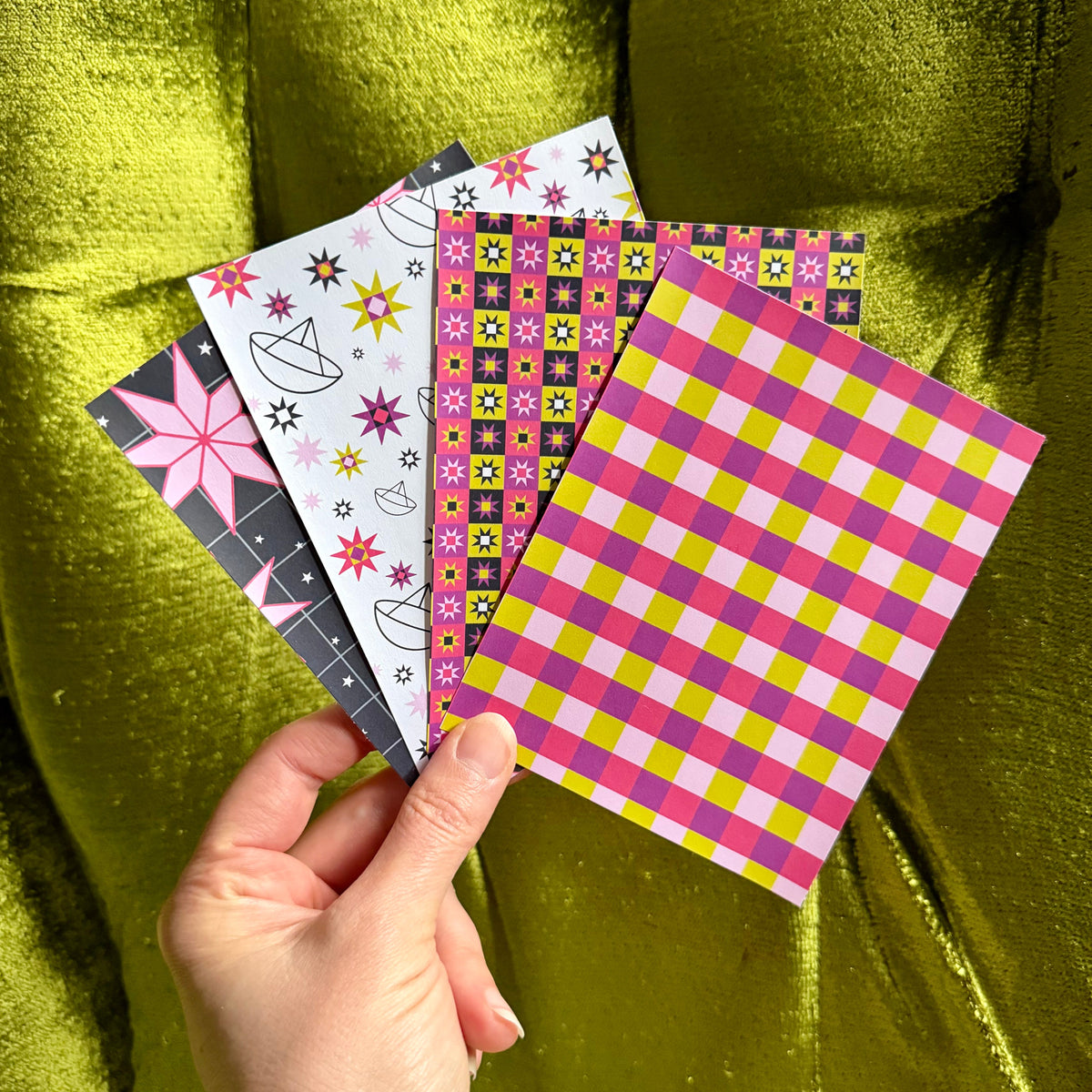 A hand holds four patterned sticker sheets against a shiny olive-green velvet background. The cards feature various colorful geometric and floral designs, including pink, yellow, black, white, and gray patterns.