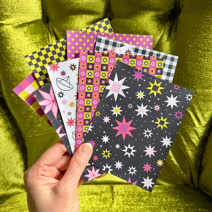 A hand holds a fan of colorful patterned sticker sheets against a shiny olive-green velvet chair. The designs include stars, stripes, polka dots, and geometric patterns in pink, yellow, black, white, and gray.