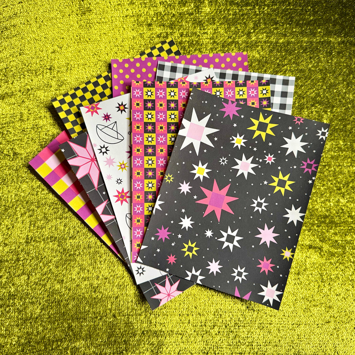 A fan of colorful patterned sticker paper sheets arranged on a bright yellow textured background. The papers feature various designs including star prints, checkered patterns, polka dots, and abstract shapes in pink, yellow, black, white, and gray.