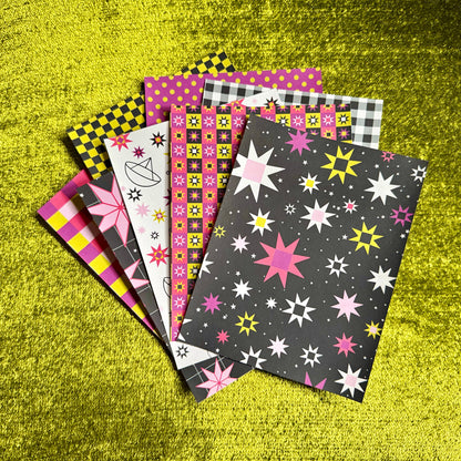 A fan of colorful patterned sticker paper sheets arranged on a bright yellow textured background. The papers feature various designs including star prints, checkered patterns, polka dots, and abstract shapes in pink, yellow, black, white, and gray.