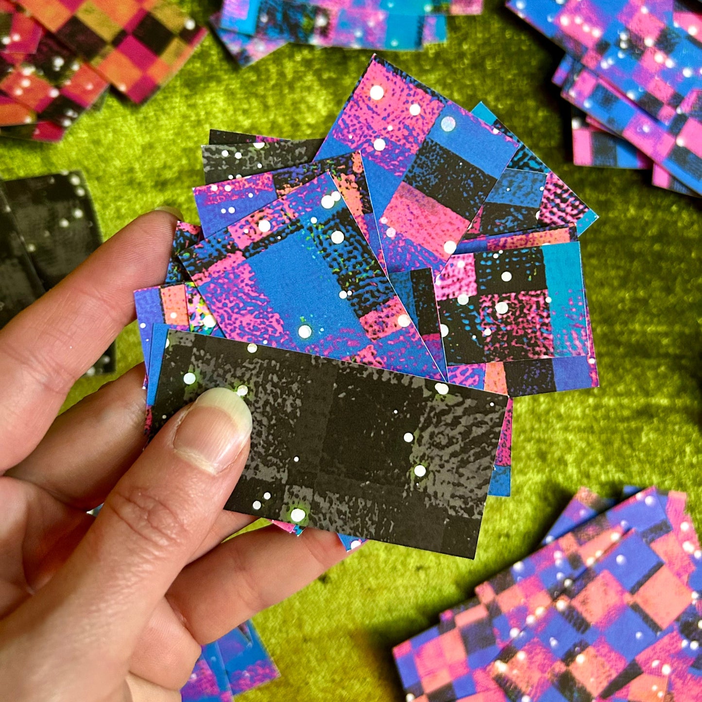 Hand holding a colorful, patterned sticker sheet with more stickers in the background on a green surface