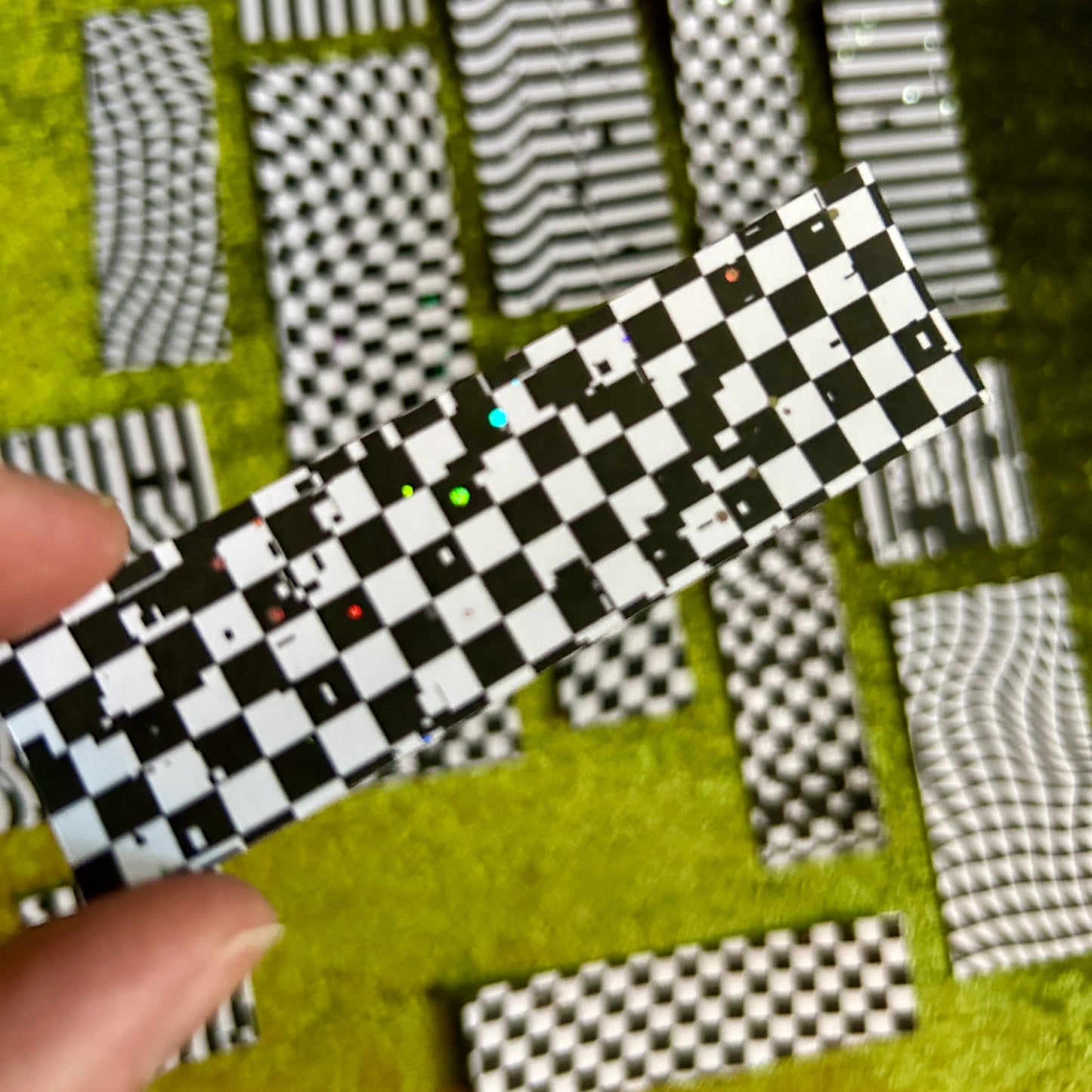Black and white checkered rectangular sticker scraps with holographic sparkles on a green textured surface