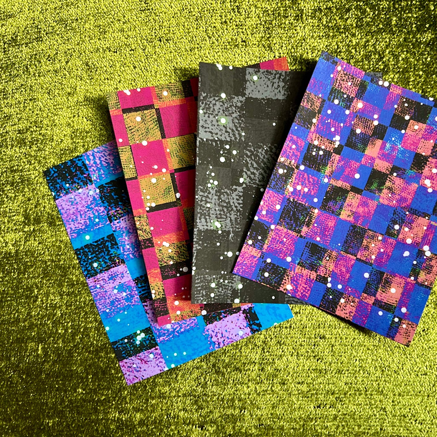 Four colorful checkered papers with glittery patterns on a green textured surface