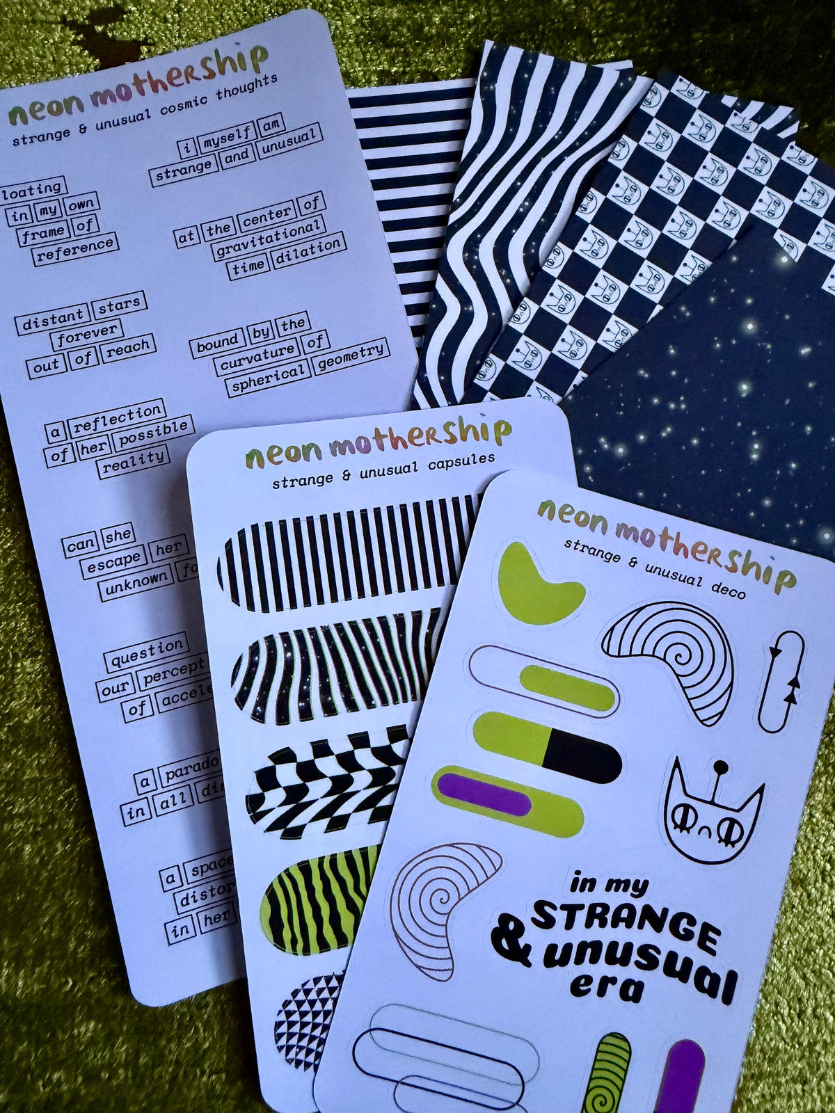 Three printed sticker sheets with abstract black, white, green, and purple shapes and text, arranged on a textured gold background.