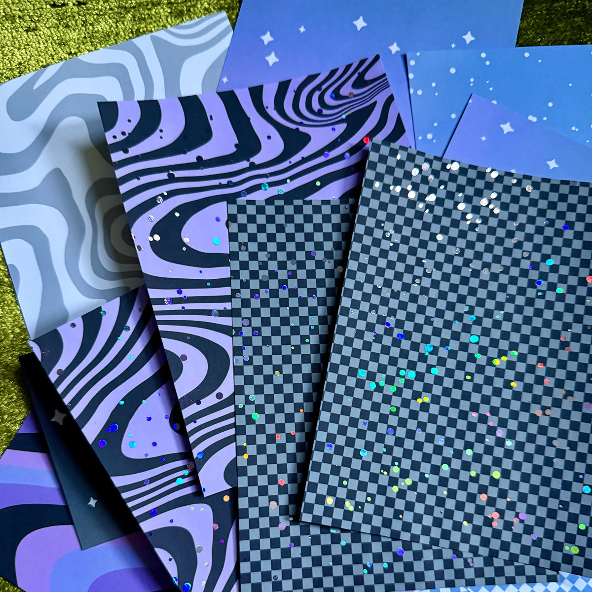 A fan arrangement of patterned paper sheets in shades of blue, purple, black, and white, featuring designs like wavy stripes, checkerboards, stars, and holographic confetti, displayed on a textured green surface.