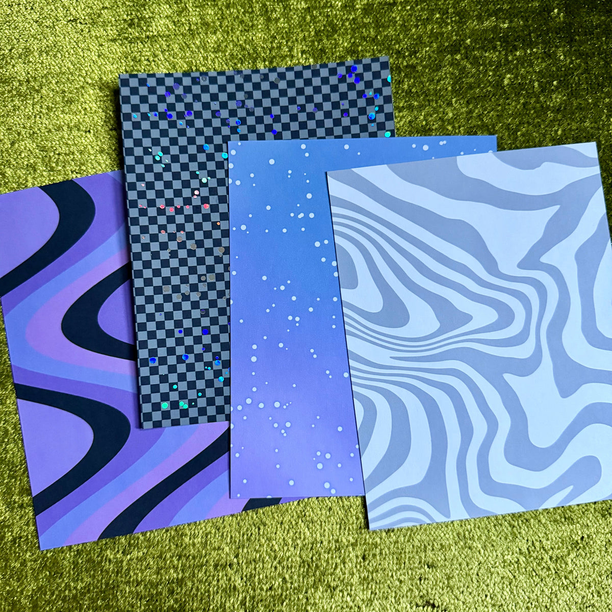 Four patterned art cards or sheets fanned out on a textured green surface. The designs include purple and black wavy lines, a black and gray checkerboard with multicolor dots, a blue-to-purple gradient with white speckles, and a light blue and white wavy pattern.