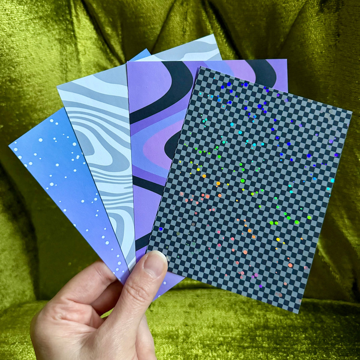 A hand holds a fan of four patterned art cards or prints against a shiny olive-green velvet background. The cards feature abstract designs in shades of blue, purple, black, and white, including splatter, wavy line, and checkerboard patterns.