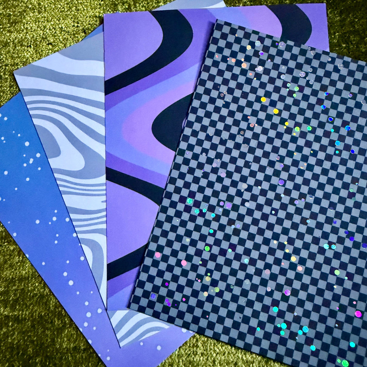 Four patterned paper sheets or cards fanned out on a textured gold surface. The designs include white stars on blue, lavender and purple wavy lines, and a black-and-gray checkerboard with small iridescent dots.