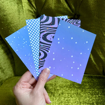 A hand holds four patterned art cards or prints fanned out against a shiny olive-green velvet chair. The cards feature space-themed designs including a blue-to-purple gradient with white stars, a black and purple wavy pattern, a blue-and-white checkerboard print, and a purple-and-black zebra-like pattern.