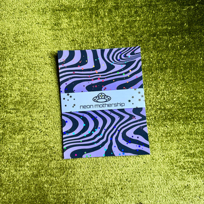 A rectangular product with a purple and black wavy zebra-like pattern and small multicolor confetti dots, wrapped with a white paper band reading 'neon mothership' and a small crown logo, placed on a textured lime-green surface.