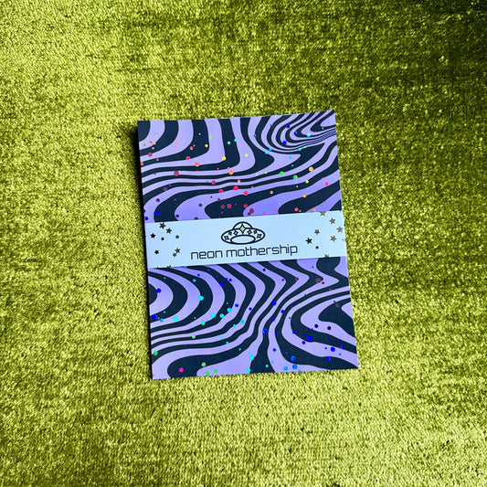 A rectangular product with a purple and black wavy zebra-like pattern and small multicolor confetti dots, wrapped with a white paper band reading 'neon mothership' and a small crown logo, placed on a textured lime-green surface.