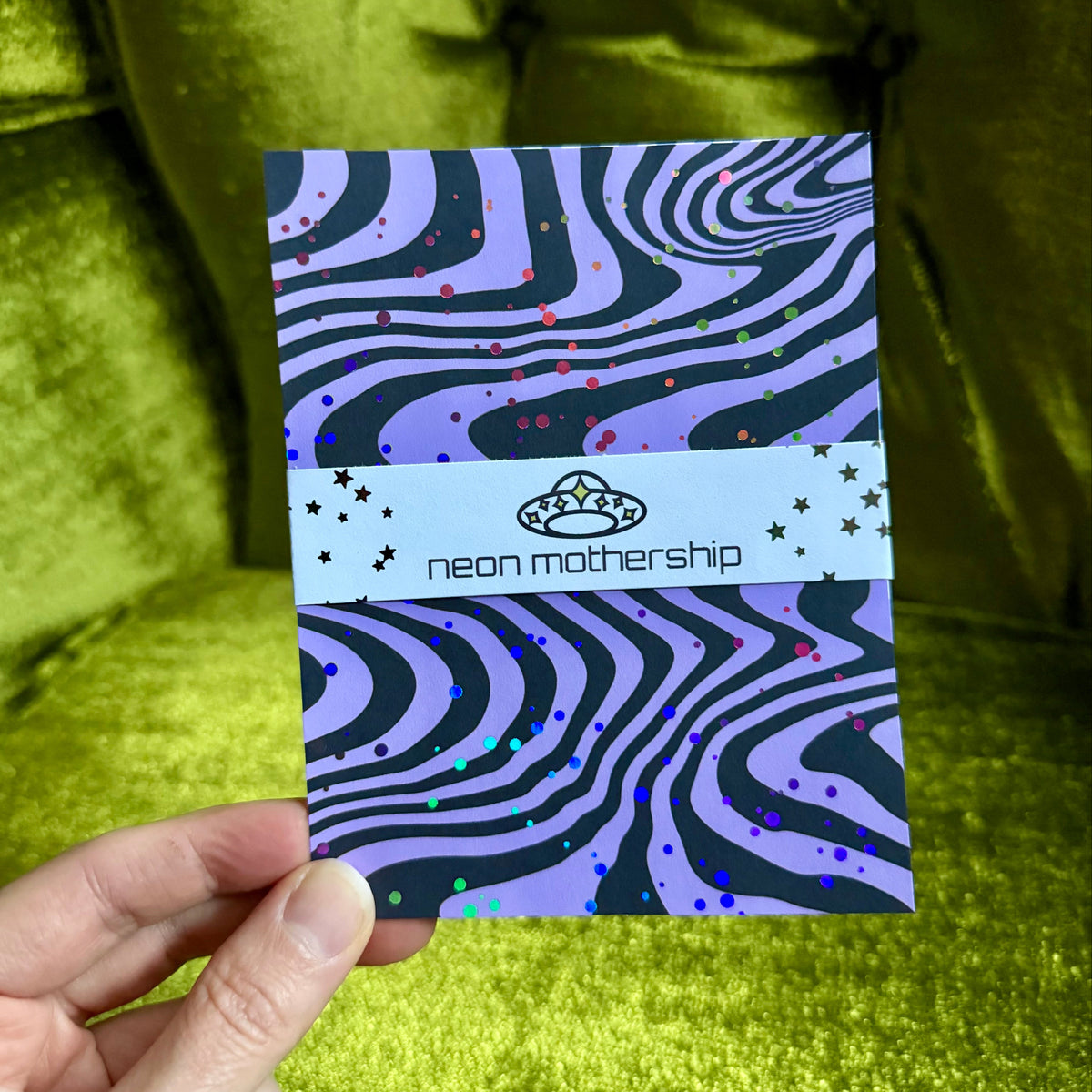 A hand holds a rectangular card featuring a purple and black wavy pattern with small multicolor glitter dots and a white horizontal band across the middle that reads 'neon mothership' with a UFO logo. The background is a shiny olive-green velvet surface.