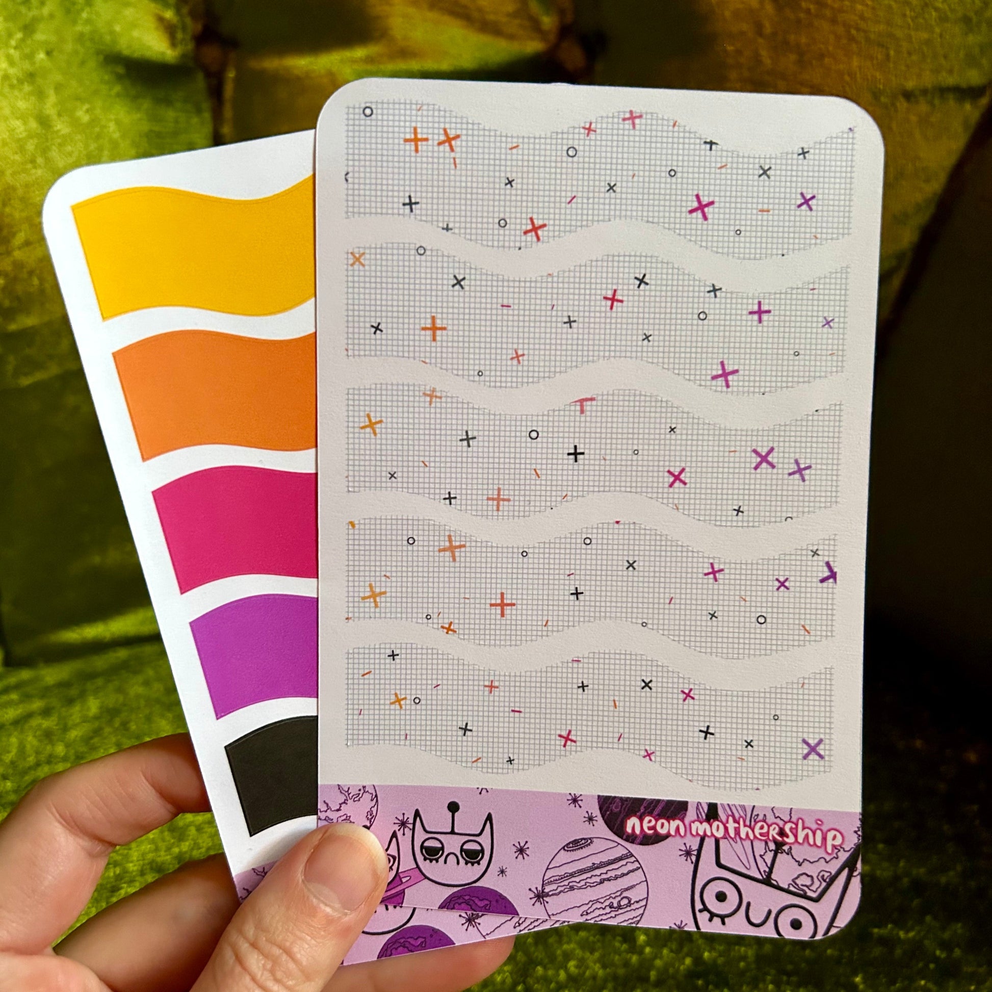 Hand holding a colorful sticker sheet with patterned design against a green fabric background