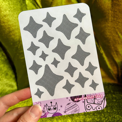 sticker sheet with black geometric patterns on a white background held by a hand against a green textured fabric background.