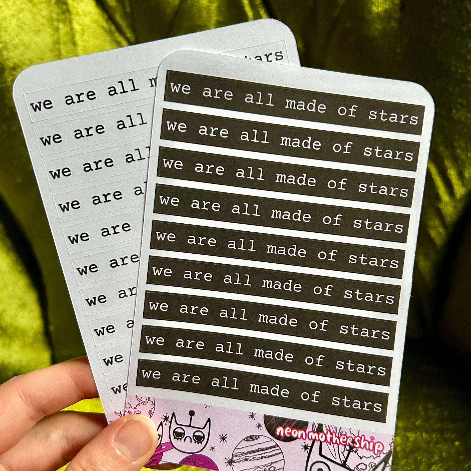 Hand holding two sticker sheets with 'we are all made of stars' text against a green fabric background
