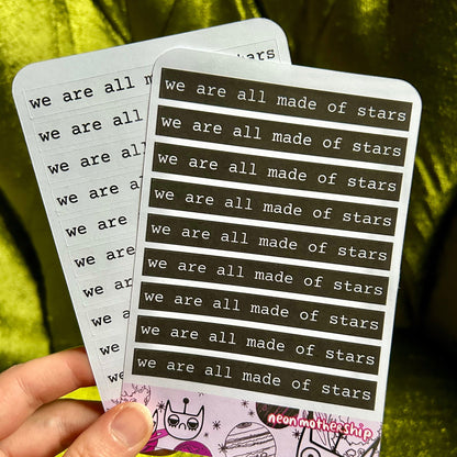 Hand holding two sticker sheets with 'we are all made of stars' text against a green fabric background