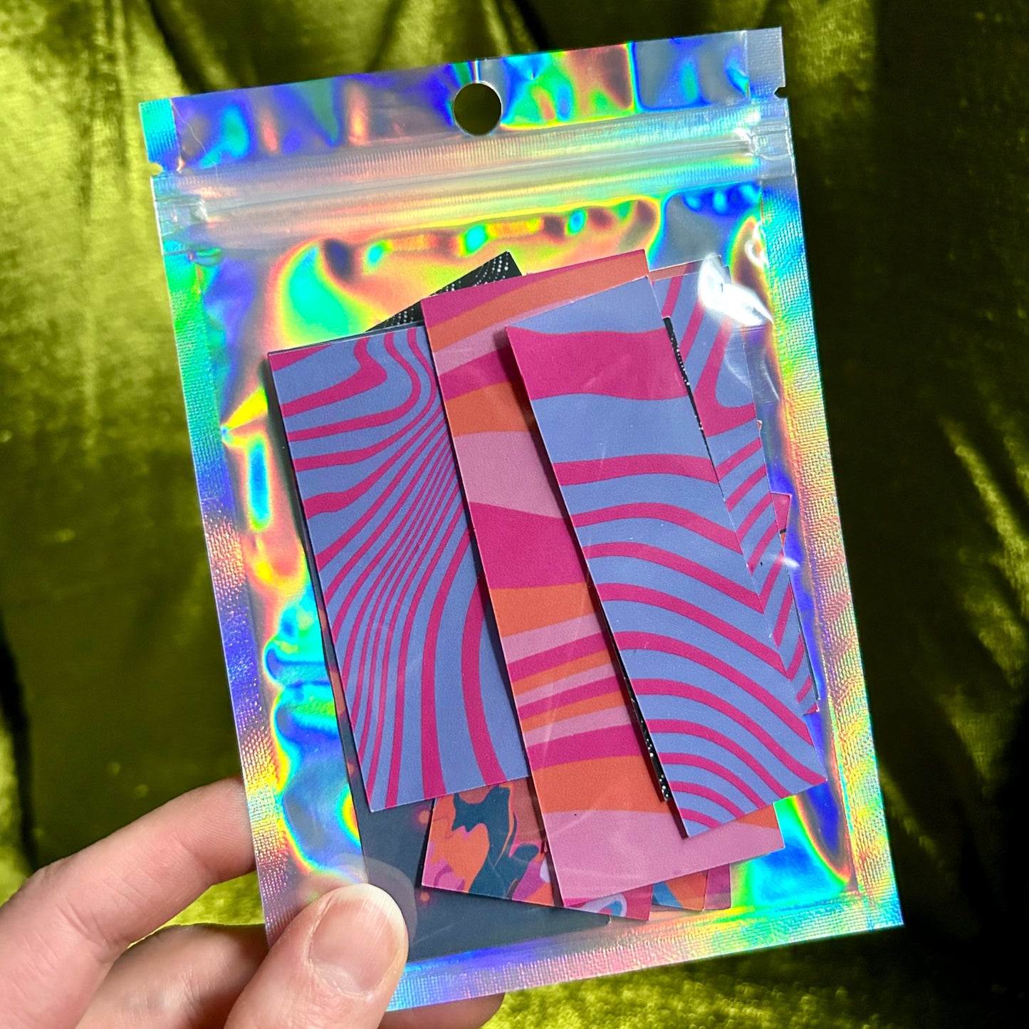 Holographic packaging with colorful striped papers held by a hand against a green fabric background