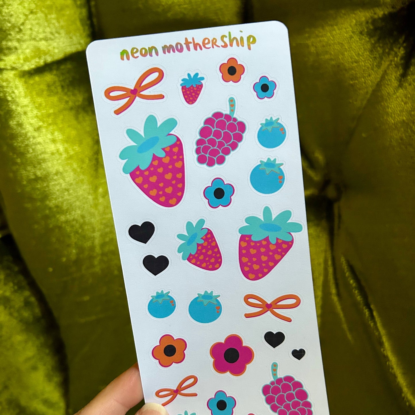 Sheet of colorful stickers with strawberries, flowers, and bows held by a hand against a green fabric background.