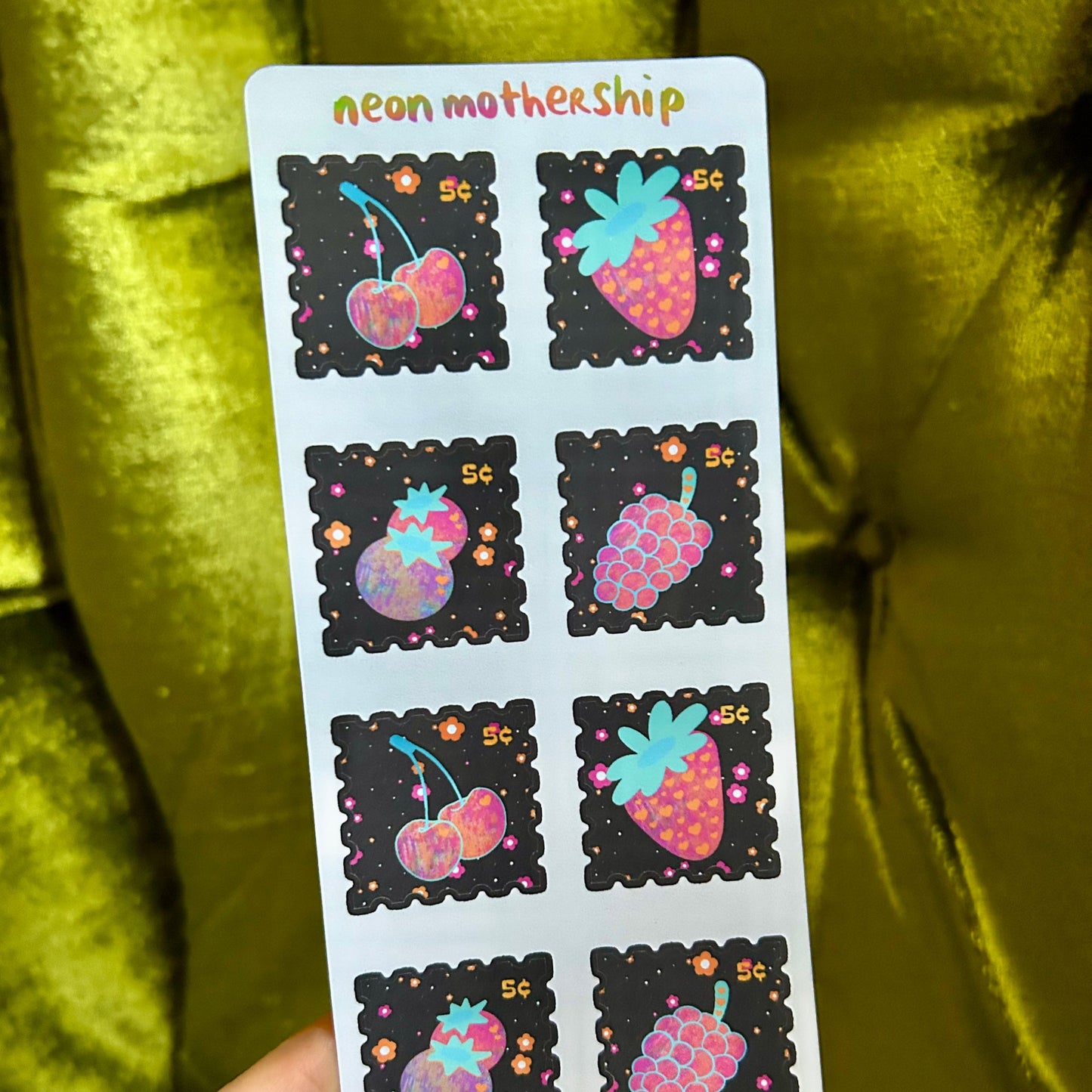 Sheet of colorful stickers with fruit designs held by a hand against a green fabric background.