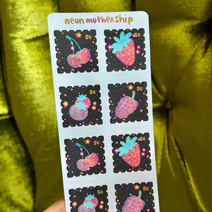 Sheet of colorful stickers with fruit designs held by a hand against a green fabric background.