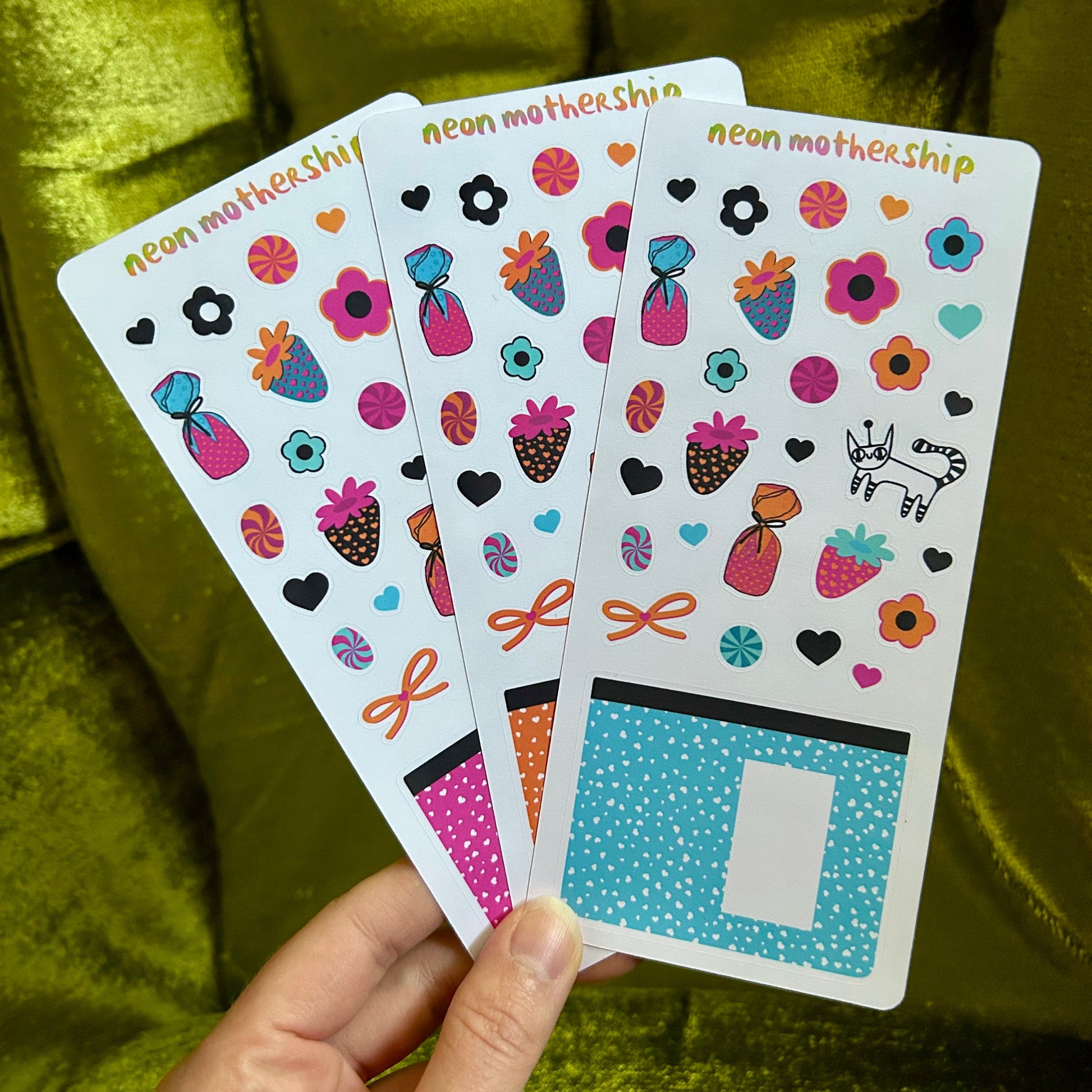 Hand holding three sheets of colorful stickers with 'neon mothership' branding against a green fabric background.
