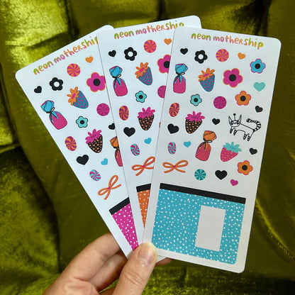 Hand holding three sheets of colorful stickers with 'neon mothership' branding against a green fabric background.
