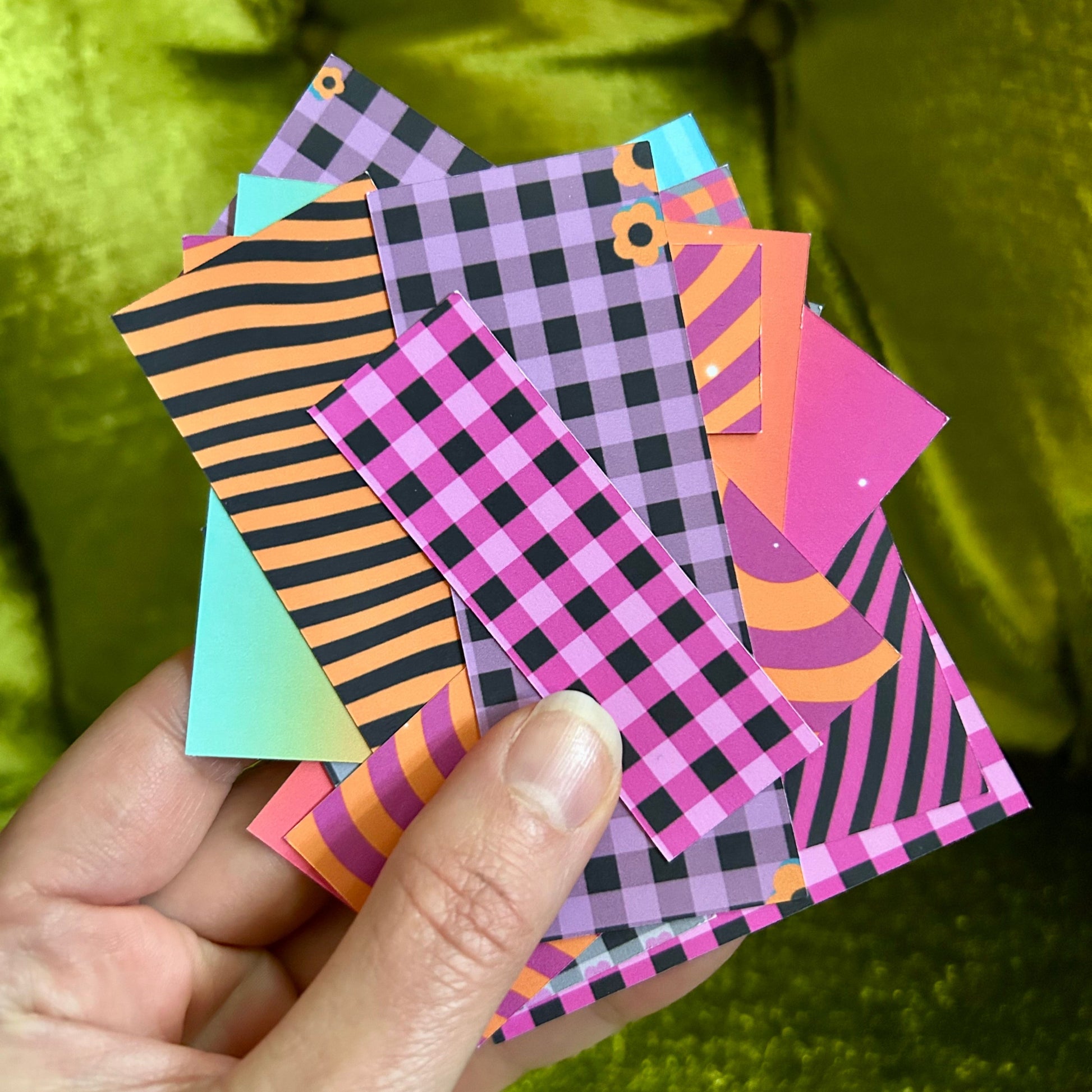 Hand holding a stack of colorful patterned sticker paper sheets against a green background