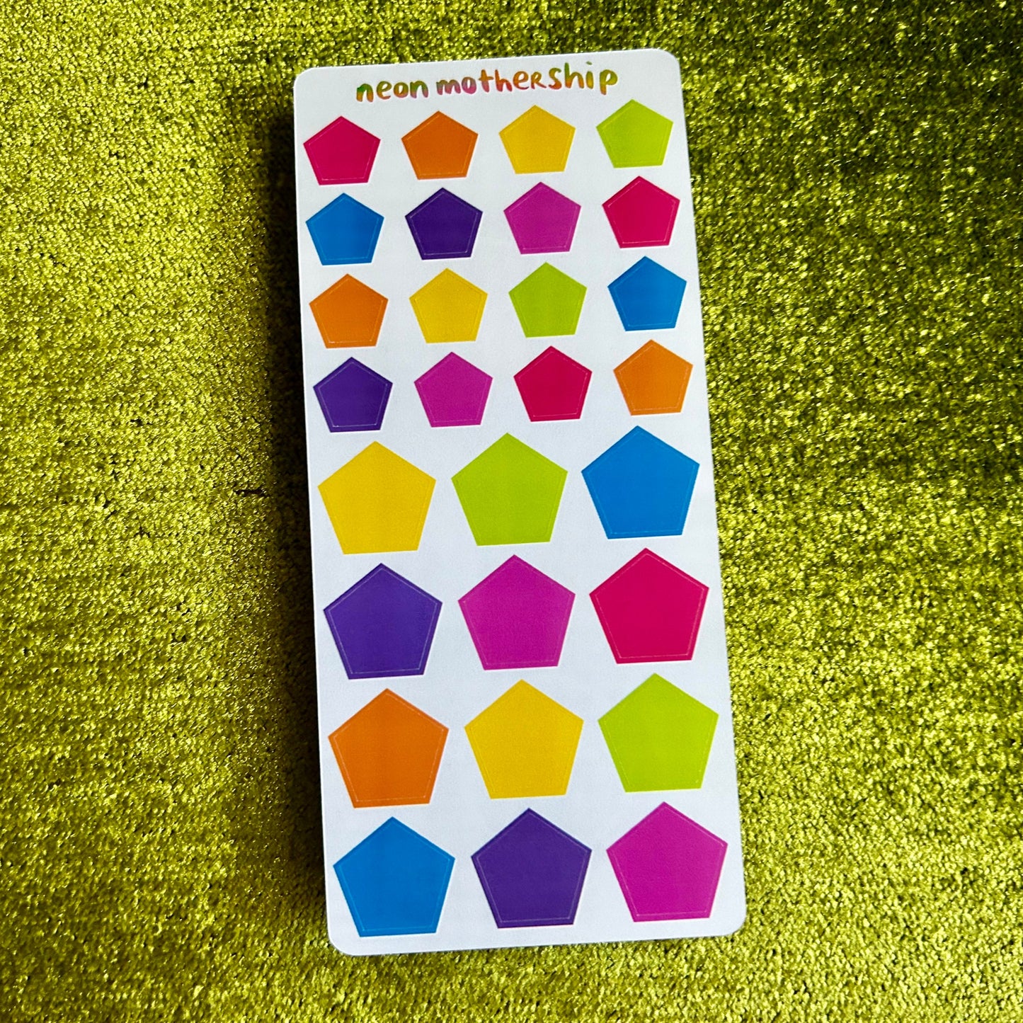 Colorful pentagon-shaped stickers on a white card with 'neon motherhip' branding, placed on a green textured surface.