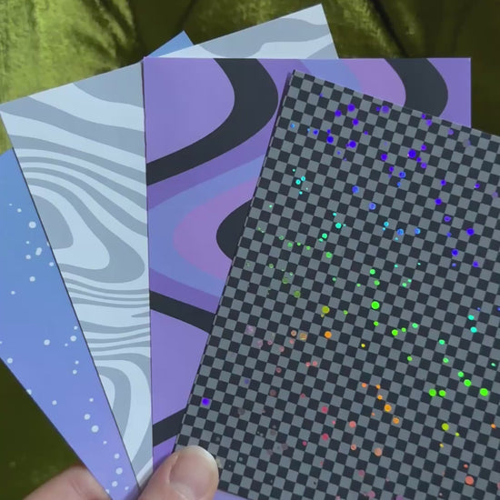 a hand holds four patterned sticker sheets and waves the sheets back and forth to catch the light on holographic sparkles